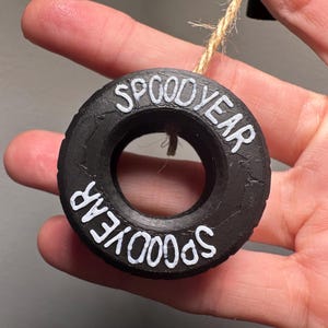 May include: Black, circular ornament with the words "SPOOD YEAR" in white lettering. The ornament is made to look like a tire and is suspended by a natural twine string.