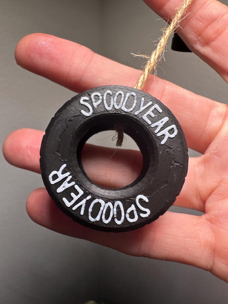 May include: Black, circular ornament with the words "SPOOD YEAR" in white lettering. The ornament is made to look like a tire and is suspended by a natural twine string.