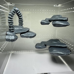 Op de afbeelding: Grijze terrariumdecoratie van hars, inclusief een rotsboog en verschillende gelaagde rotsformaties. De stukken zijn ontworpen om een naturalistische omgeving te creëren in een reptielenverblijf. De decoratie wordt getoond in een transparante behuizing.