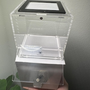 May include: A clear acrylic terrarium with a black mesh top and a white base. The terrarium has a hinged door and a small, clear compartment inside. The brand name "Heather's Nightmare" is visible on the front.