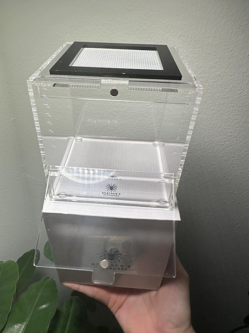 May include: A clear acrylic terrarium with a black mesh top and a white base. The terrarium has a hinged door and a small, clear compartment inside. The brand name "Heather's Nightmare" is visible on the front.