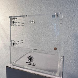 May include: A clear acrylic terrarium with a hinged lid and ventilation holes. The front of the terrarium features the text "Heather's Hideaways" with a spider graphic. The terrarium is designed for small pets.