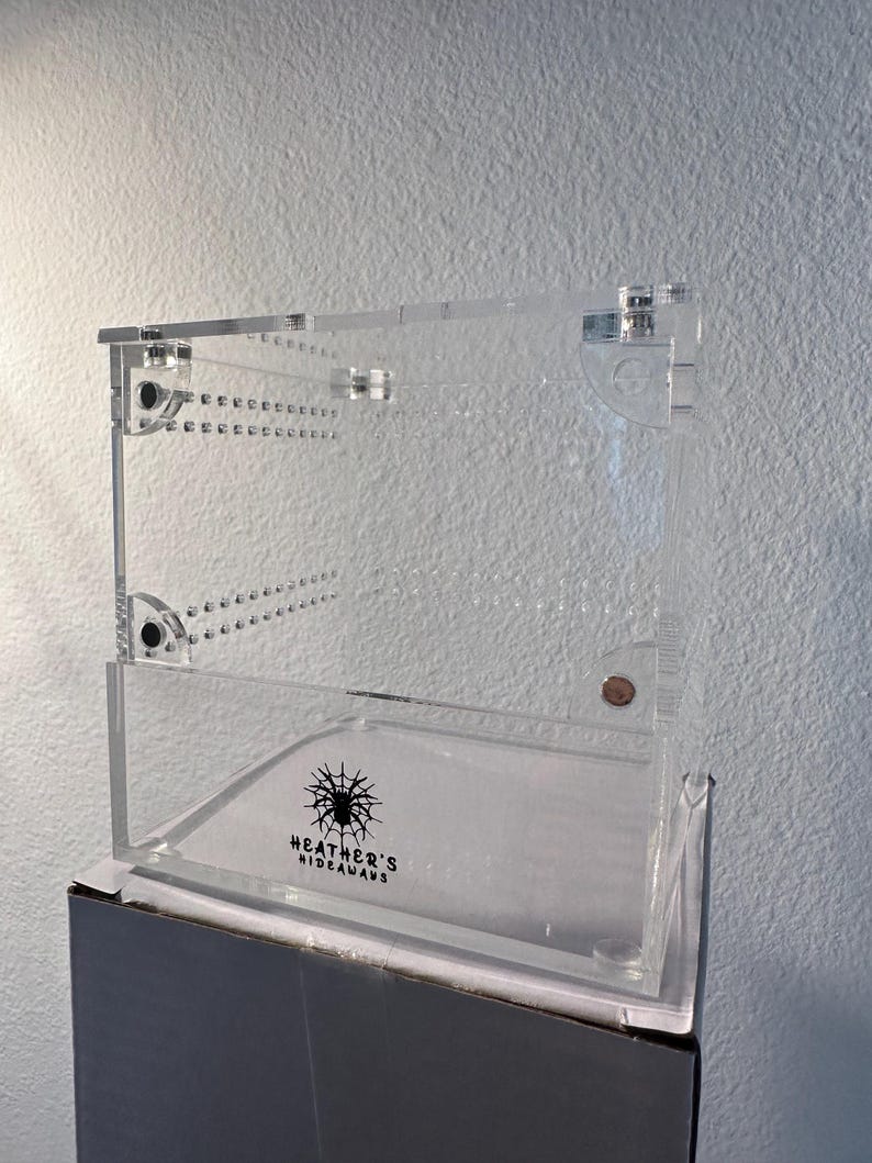 May include: A clear acrylic terrarium with a hinged lid and ventilation holes. The front of the terrarium features the text "Heather's Hideaways" with a spider graphic. The terrarium is designed for small pets.