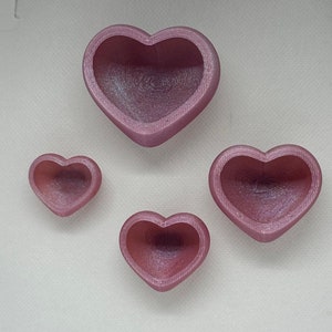 May include: Four pink heart-shaped bowls of varying sizes. The bowls are made of a smooth, shiny material and have a slightly iridescent finish.
