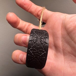 May include: A black, cylindrical ornament with a spiderweb design. The ornament is suspended by a natural twine string. The object is held in a hand against a neutral background.