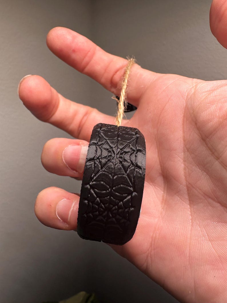 May include: A black, cylindrical ornament with a spiderweb design. The ornament is suspended by a natural twine string. The object is held in a hand against a neutral background.