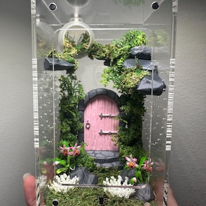 May include: A clear acrylic terrarium with a miniature fairy garden scene. The scene includes a pink door, moss, rocks, and artificial plants. The terrarium has a small, clear, round container at the top.