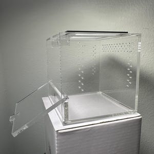 May include: A clear acrylic terrarium with a hinged door and ventilation holes. The open door reveals the interior, and the terrarium sits atop a white cardboard box. The design is simple and functional, suitable for housing small animals.