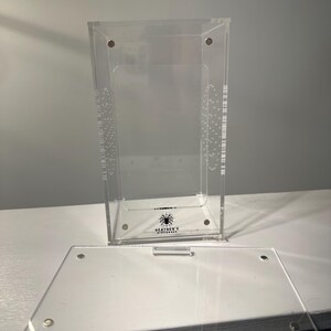 May include: A clear acrylic terrarium with a removable lid. The terrarium has a rectangular shape with a logo that reads "Heather's Hideaways" and a spider graphic. The lid is off to the side.