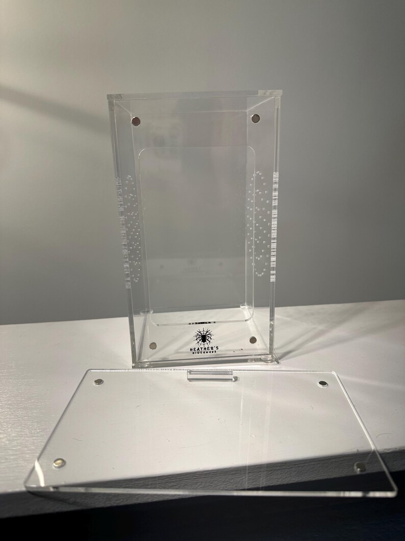 May include: A clear acrylic terrarium with a removable lid. The terrarium has a rectangular shape with a logo that reads "Heather's Hideaways" and a spider graphic. The lid is off to the side.