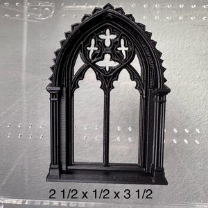 May include: Black gothic window with an arched top and cross-shaped details. The window measures 6.35 cm high, 1.27 cm wide, and 8.89 cm long. The window is a decorative element.