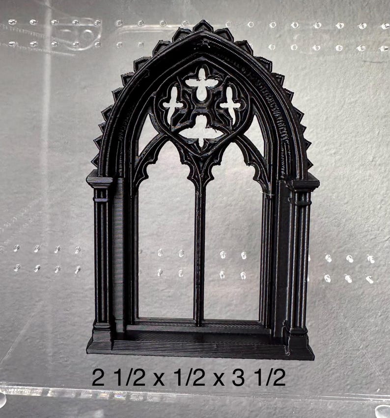 May include: Black gothic window with an arched top and cross-shaped details. The window measures 6.35 cm high, 1.27 cm wide, and 8.89 cm long. The window is a decorative element.