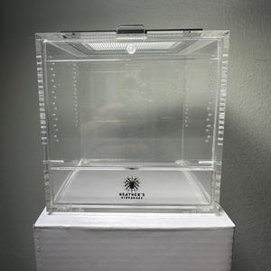 May include: A clear acrylic terrarium with a hinged lid and ventilation. The terrarium sits on a white box. The text "HEATHER'S HIDEAWAYS" is printed on the front of the terrarium. The terrarium is designed for small pets.