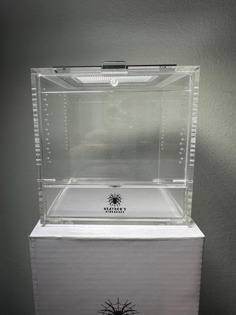 May include: A clear acrylic terrarium with a hinged lid and ventilation. The terrarium sits on a white box. The text "HEATHER'S HIDEAWAYS" is printed on the front of the terrarium. The terrarium is designed for small pets.