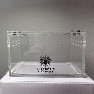 Puede incluir: Un terrario de acrílico transparente con forma rectangular. La parte delantera del terrario tiene pequeños agujeros y una tapa con bisagras. Las palabras "HEATHER'S HIDEAWAYS" están impresas en negro con un gráfico de telaraña. El terrario está sobre una caja blanca.