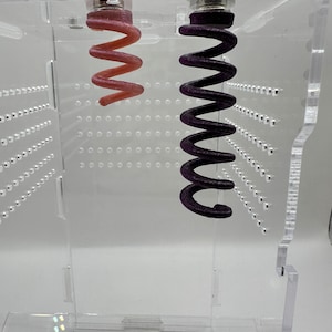 May include: Two coiled springs hanging from the top of a clear acrylic box. The spring on the left is pink and the spring on the right is purple.
