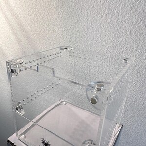 May include: A clear acrylic terrarium with a rectangular shape. The enclosure features multiple drilled holes and is held together with clear fasteners. The base has a white surface. The text "HEATHER'S HIDEAWAYS" is visible.