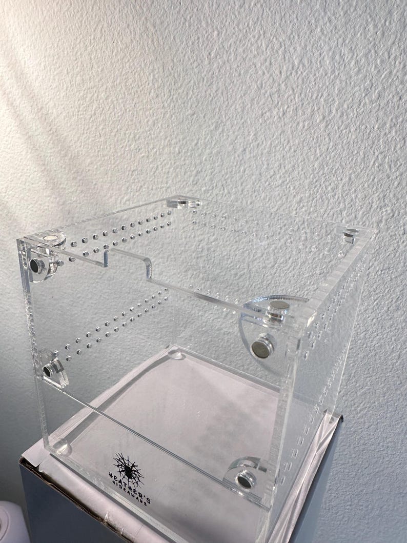 May include: A clear acrylic terrarium with a rectangular shape. The enclosure features multiple drilled holes and is held together with clear fasteners. The base has a white surface. The text "HEATHER'S HIDEAWAYS" is visible.