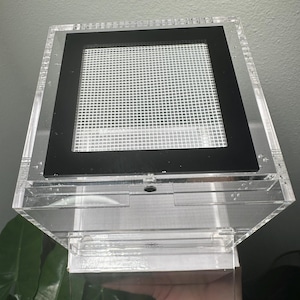 May include: A clear acrylic terrarium with a black framed mesh top. The terrarium has a rectangular shape and is designed to house small animals or insects. The design includes a hinged lid and a clear front panel for viewing.