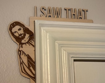May include: Wooden door frame decoration featuring a laser-cut image of Jesus peeking around the corner. The text "I SAW THAT" is above the image. The decoration is made of light-colored wood and is designed to be placed on a door frame.