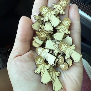 May include: A handful of small, wooden figurines of Jesus. Each figurine is light brown with a darker brown outline, depicting Jesus with a beard and long hair, wearing a robe. The figurines are held in a person's hand.