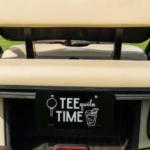 May include: A black license plate on a golf cart with the words "TEEquila TIME" in white. The plate features a golf ball and tee graphic, and a cocktail graphic. The golf cart is beige.
