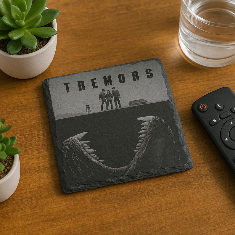 May include: A square Tremors coaster made of dark gray slate. The coaster features the movie title and a graphic of the characters and a monster's jaws. A remote, a glass of water, and a succulent plant are in the background.