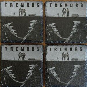 May include: Four slate coasters featuring the movie title "Tremors". Each coaster has a gray top section with the movie title and figures, and a black bottom section with a monster's jaws. The coasters are square.