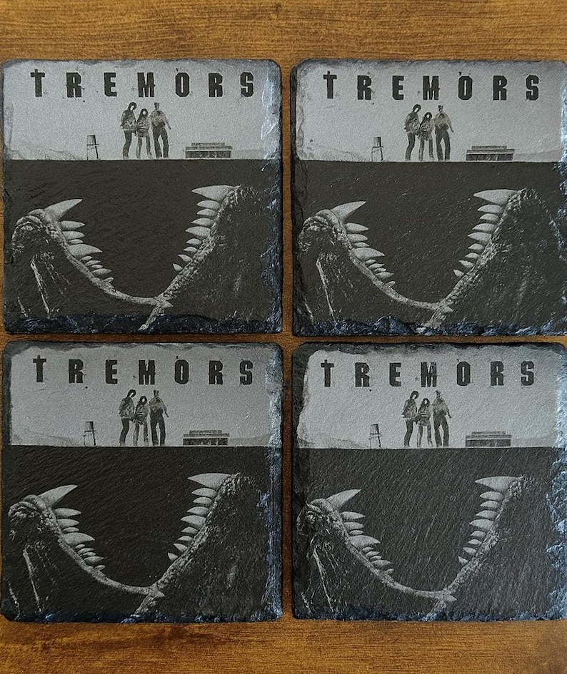 May include: Four slate coasters featuring the movie title "Tremors". Each coaster has a gray top section with the movie title and figures, and a black bottom section with a monster's jaws. The coasters are square.