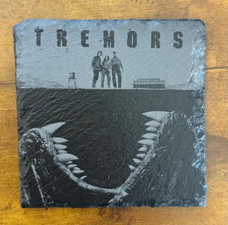May include: Square slate coaster featuring the word "TREMORS" above a graphic of three people and a monster's open jaws. The coaster is dark gray with a textured surface, suitable for drinks.