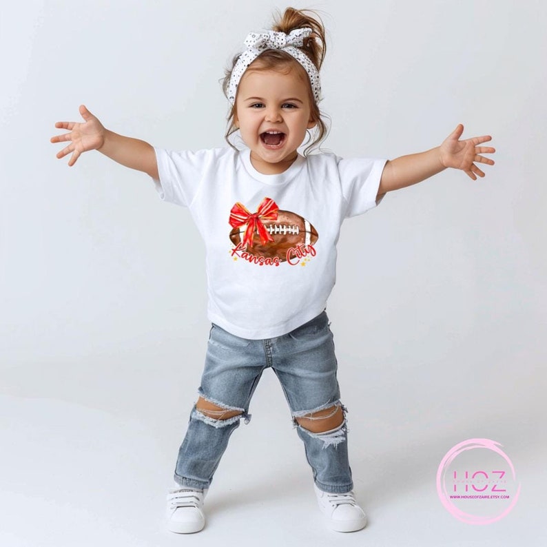 May include: A white t-shirt featuring a brown and red football with a red bow on top. The football has the words "Kansas City" written on it. A young child is wearing the shirt and smiling.