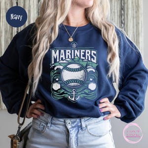 May include: Navy blue sweatshirt featuring a baseball design with the word "MARINERS" in white and green. The graphic includes a baseball, waves, and an anchor. The sweatshirt is a crew neck style.