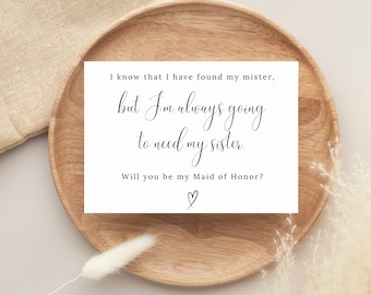 May include: A white card with black text on a wooden plate. The text reads: "I know that I have found my mister, but I'm always going to need my sister. Will you be my Maid of Honor?" with a heart symbol at the bottom.
