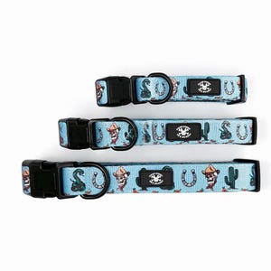 May include: Three dog collars with a light blue background and a repeating pattern of cowboy-themed illustrations. The collars have black plastic buckles and metal D-rings. The pattern includes cacti, snakes, horseshoes, and cowboy skulls.