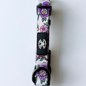 May include: A white pet collar featuring a floral pattern with purple and pink flowers, green leaves, and brown stems. The collar includes black plastic hardware, a black D-ring, and a black rectangular tag with the text Tuff Bones Pet Stuff.