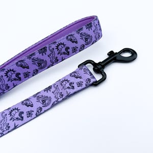 May include: A lavender dog leash featuring a repeating black line art pattern of skulls, butterflies, dice, and feline figures. The leash includes a black metal swivel snap hook and has a solid lavender interior lining on the handle loop.