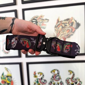 May include: A dark gray pet collar featuring a repeating pattern of skulls, daggers, and green and red snakes. The collar includes a black metal quick-release buckle and a black metal D-ring for attaching a leash, constructed from durable webbing.