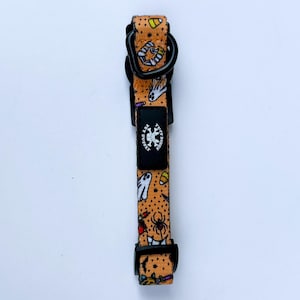 May include: An orange pet collar featuring a repeating Halloween pattern of ghosts, spiders, candy corn, and bats on a black polka dot background. The collar includes a black plastic buckle, a black metal D-ring, and a black rectangular tag with a white skull logo and the text Tuff Bonez Pet Supply.