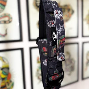 May include: A black nylon dog collar featuring a repeating pattern of skulls, roses, and barbed wire. The collar includes a black metal D-ring, a heavy-duty buckle, and a black fabric tag with the white text TUFF BONES and a skull logo.