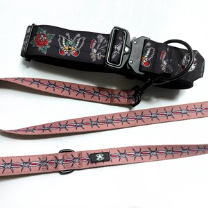 May include: A black dog collar featuring a colorful tattoo-style print of roses and butterflies with a black metal buckle, paired with a dusty pink leash decorated with a repeating pattern of silver and red barbed wire and a black metal D-ring.
