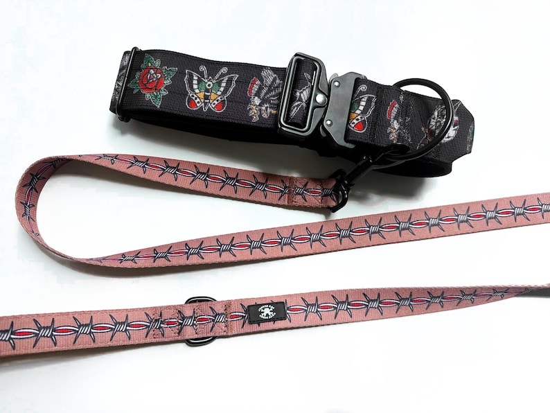 May include: A black dog collar featuring a colorful tattoo-style print of roses and butterflies with a black metal buckle, paired with a dusty pink leash decorated with a repeating pattern of silver and red barbed wire and a black metal D-ring.