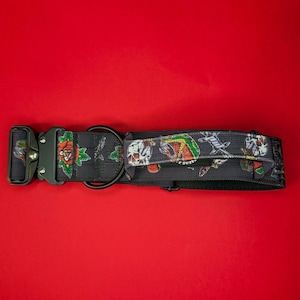 May include: Black dog collar with a repeating pattern of roses, skulls, and other tattoo-style graphics. The collar has a black metal buckle and a D-ring for leash attachment. The background is a solid red.