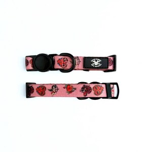 May include: Two pink dog collars with black hardware. The collars feature a repeating pattern of tattoo-style illustrations, including hearts, cherubs, and anchors. One collar has a black label with white text and a skull logo.