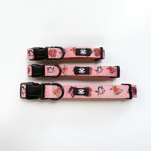 May include: Three pink pet collars of varying widths featuring a repeating tattoo-style print of cupids, hearts, and panthers. Each collar includes a black plastic side-release buckle, a black metal D-ring, and a black rectangular brand label.