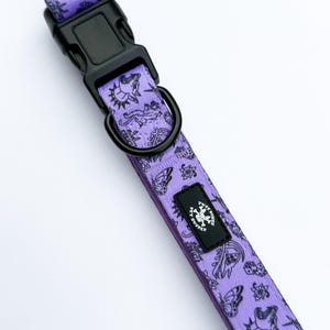 May include: A purple pet collar featuring a repeating black tattoo-style print of skulls, butterflies, dice, and wolves. The collar includes a black plastic side-release buckle, a black metal D-ring, and a black rectangular label with the text TUFF BONES.