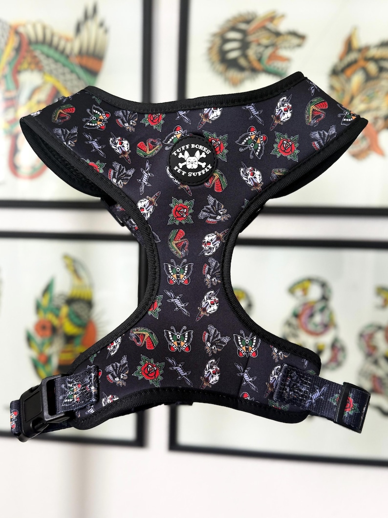 May include: Black dog harness with a tattoo-inspired print featuring roses, butterflies, skulls, and daggers. The harness has a black adjustable strap with a buckle and the text "Tuff Bones Pet Supplies" on a circular patch.