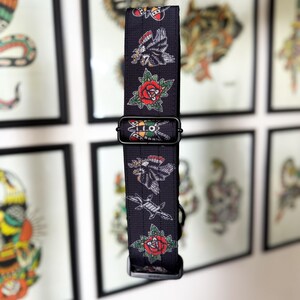 May include: A black nylon webbing strap featuring a repeating pattern of traditional tattoo-style illustrations, including red roses, eagles, and moths. The strap includes a black plastic adjustable slider and a black plastic buckle fastener.