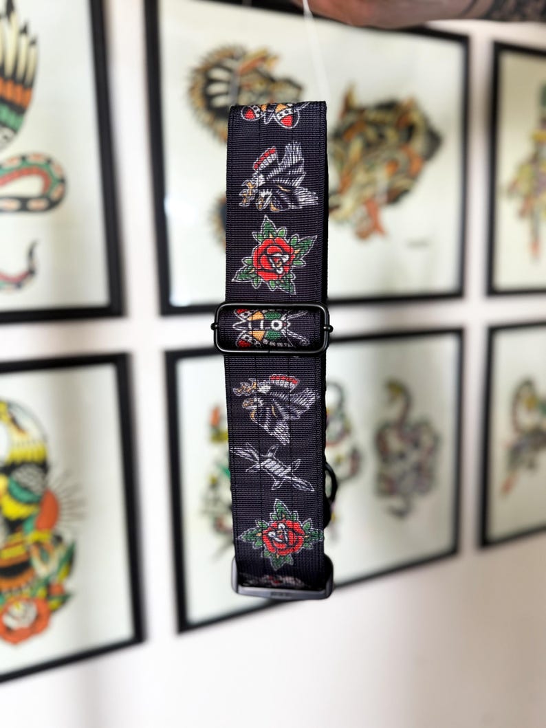May include: A black nylon webbing strap featuring a repeating pattern of traditional tattoo-style illustrations, including red roses, eagles, and moths. The strap includes a black plastic adjustable slider and a black plastic buckle fastener.