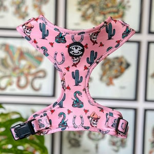 May include: Pink dog harness with a western-themed print featuring cacti, snakes, horseshoes, and skulls. The harness has a black adjustable strap and a black buckle. The harness has a patch that says "Tuffy Power Pet Supplies".