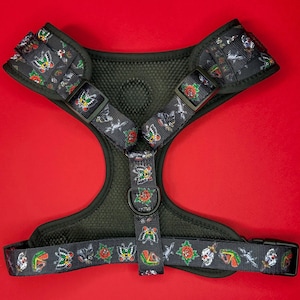 May include: A black dog harness with a colorful tattoo-inspired print featuring butterflies, roses, and skulls. The harness has a mesh lining and an adjustable buckle.